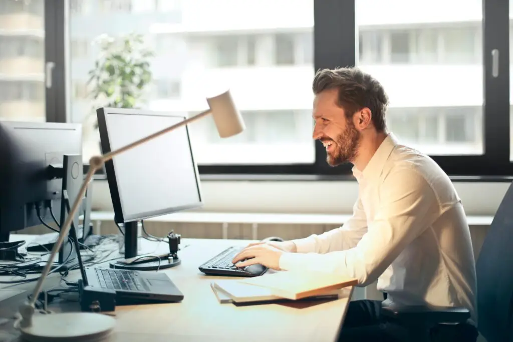 Bedrijfsafval A man smiling while working at an office desk with a computer and natural daylight streaming in through large windows.
